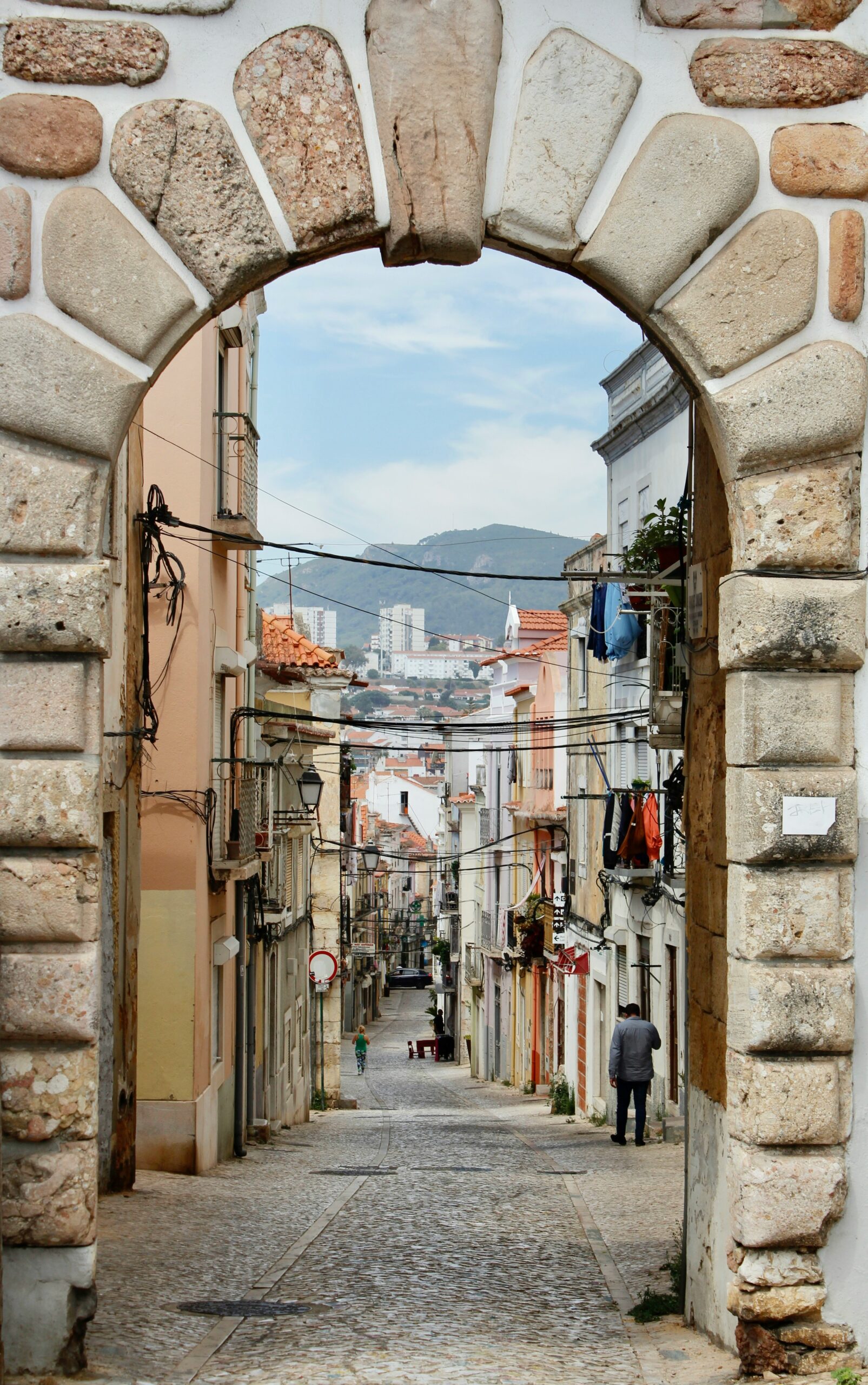 A narrow street in the historic district of Setúbal, Portugal. Buildings on the downhill stretch of a cobblestone street lined with buildings with clay tile roofs with mountains in the background.